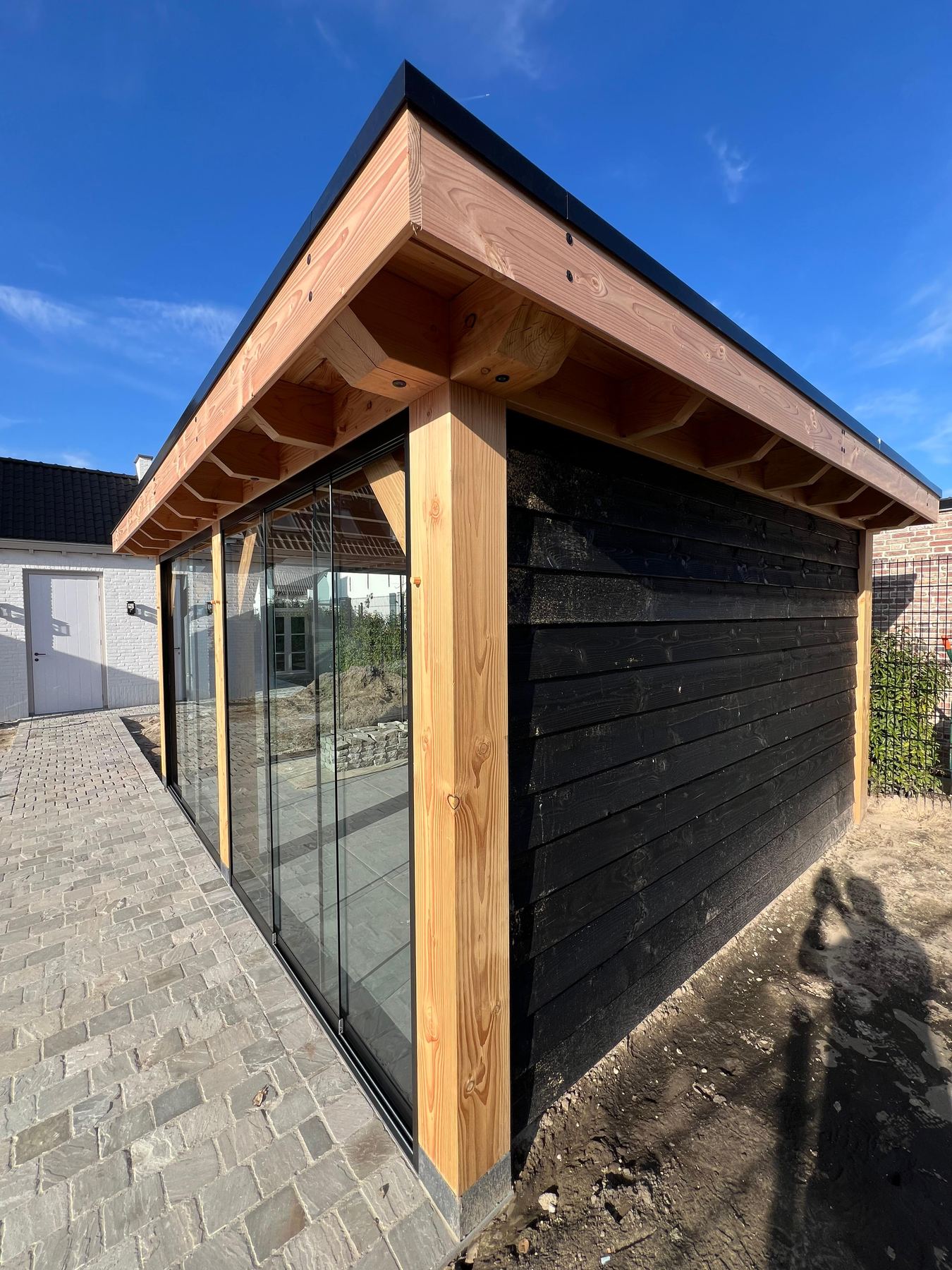Tuinkamer met glaswand en zwarte houten zijgevel onder een strakblauwe lucht