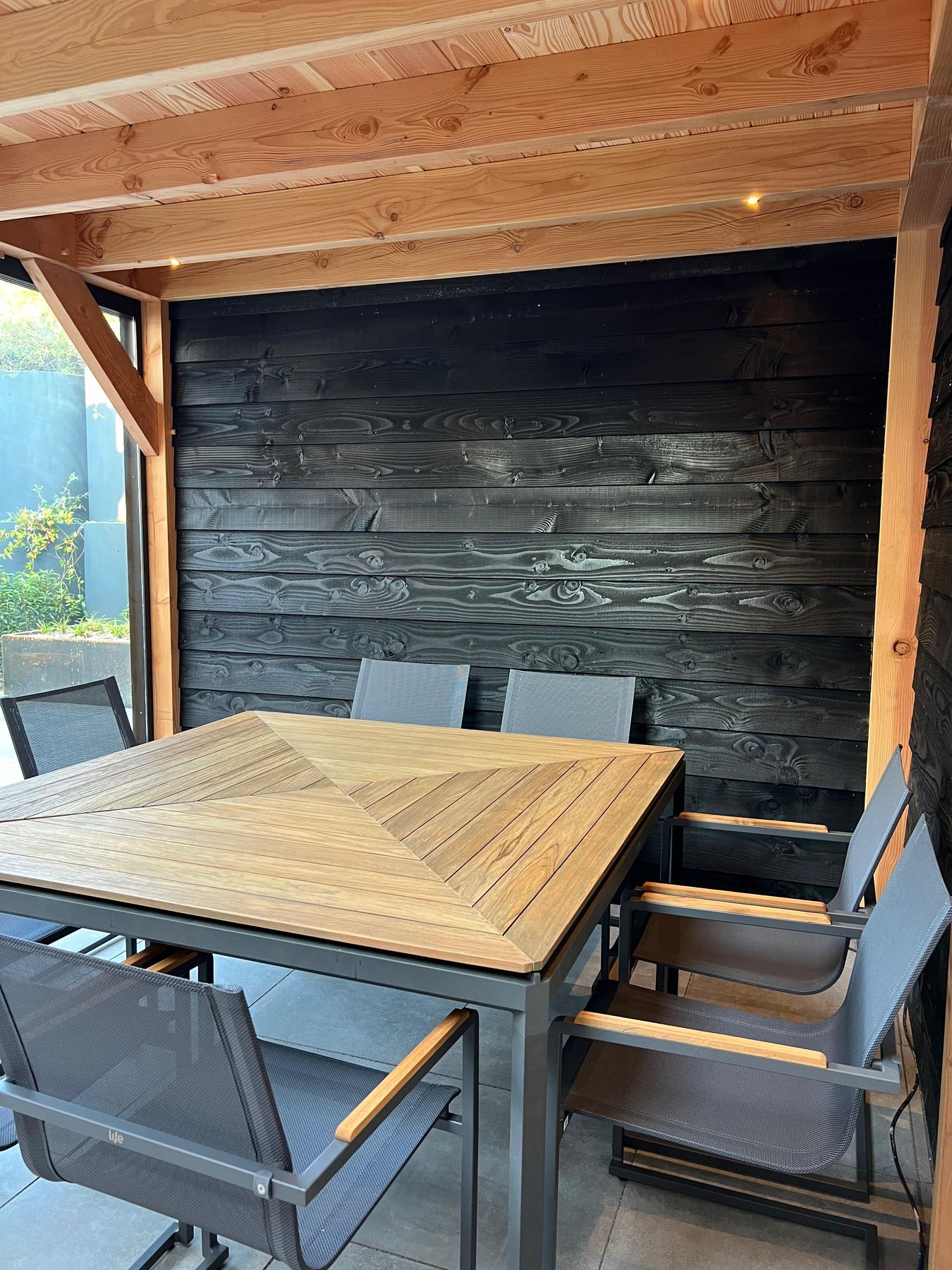 Houten eettafel met stoelen onder een overkapping tegen een zwarte houten wand